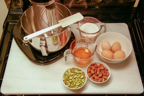 A stand mixer, a measuring cup of sugar, a measuring cup of honey, four brown eggs in a white bowl, a small white bowl of chopped pistachios, and a small white bowl of whole almonds