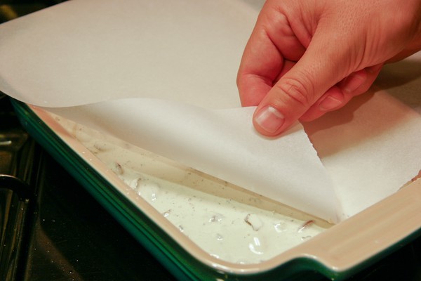 A human hand pulling back parchment paper to expose a corner of nougat in a rectangular baking dish