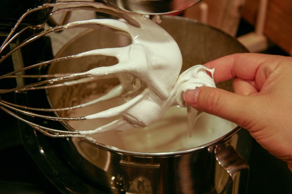 A human hand pinching something white that is on a stand mixer whisk above a stand mixer bowl, which contains something white