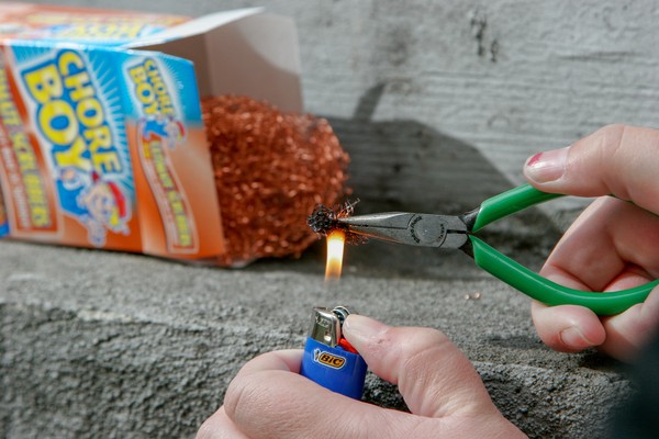 Human hand holding a piece of copper scouring pad with green-handled pliers; other hand is holding a cigarette lighter such that the flame burns the scouring pad; an open box of scouring pads is in the background