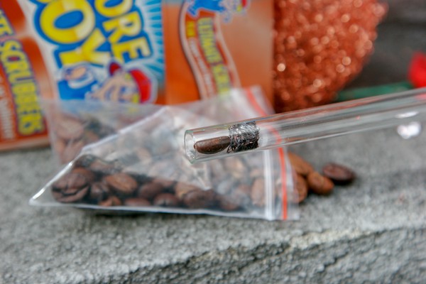 Inspecting the bean A glass tube with a piece of scouring pad and a coffee bean inside it; a bag of coffee beans is in the background along with an open box of scouring pads