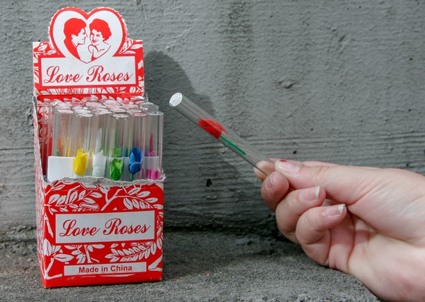 Love roses A human hand holding a glass tube with a red, plastic rose inside; an open box of multicolored love roses is in the background