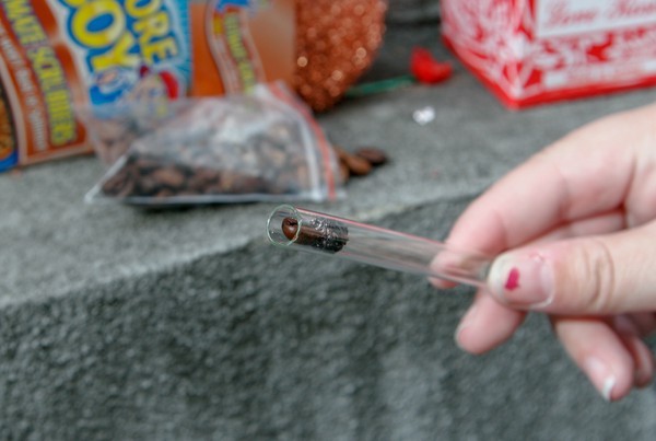 A human hand holding a glass tube with a piece of scouring pad and a coffee bean inside it; a bag of coffee beans is in the background along with open boxes of multicolored love roses and scouring pads