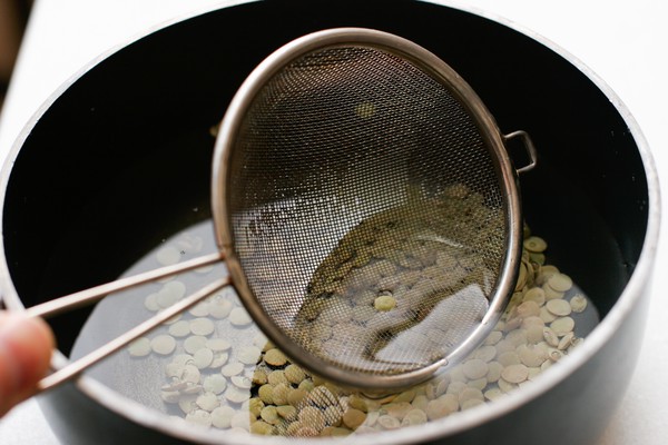 A sieve skimming a dead lentil floating on the surface of a pot of water containing cooked lentils