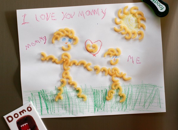 Insipid artwork A childish macaroni art drawing of an adult and a child standing on grass with the sun overhead and a heart between them; labeled, 'I love you mommy'