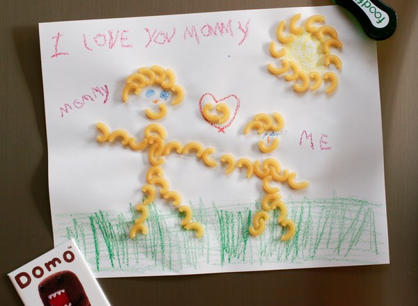 A childish macaroni art drawing of an adult and a child standing on grass with the sun overhead and a heart between them; labeled, 'I love you mommy'