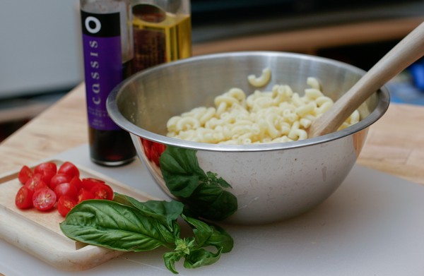 Macaroni salad ingredients Cooked macaroni in a metal bowl with a wooden spoon in it; basil and tomatoes sit on a wooden cutting board in front of the bowl; vinegar and olive oil are behind the bowl