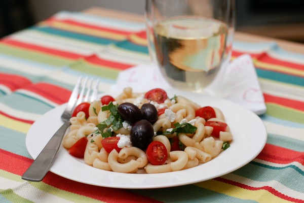 Finished macaroni salad with olives tomatoes and feta crumbles on a white plate with a metal fork; a glass of white wine is behind the plate on a napkin; all sit on a colorful tablecloth
