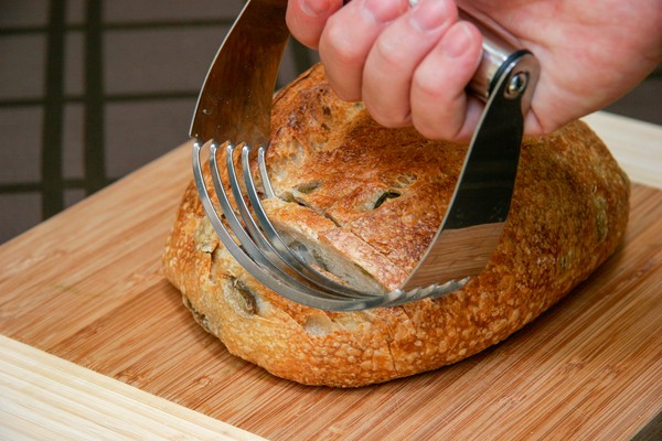 A human hand pressing a pastry blender into a loaf of sourdough bread