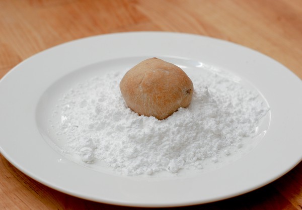 Swedish Lemon Tea Biscuit in confectioner's sugar A single, brown, potato-shaped, cookie sitting atop a mound of confectioner's sugar on a white plate