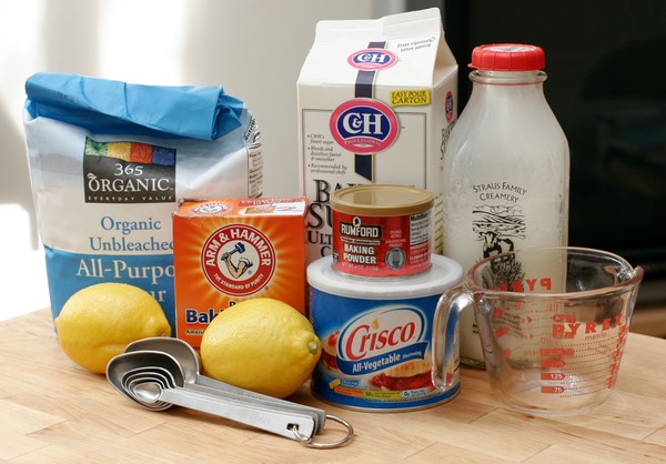 A bag of flour, a box of ultrafine sugar, a jar of milk, two lemons, a box of baking soda, a tin of baking powder, a can of Crisco, a measuring cup, and measuring spoons