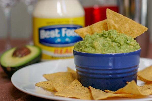 A small, blue dish of guacamole with tortilla chips in and around it on a white plate with a jar of mayonnaise and a half of an avocado in the background