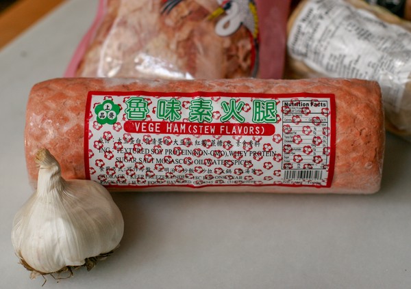 A loaf of vegetarian ham (stew flavors) with a clove of garlic in the foreground and other packaged goods in the background